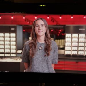 A lady with brown hair is pictured wearing a grey top with her hands behind her back. Behind her is a glasses shop with red interior and lots of LED boxes storing glasses. She is smiling.
