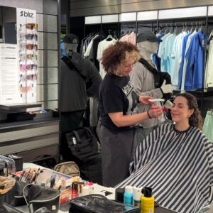 Behind the scenes filming for Ray-Ban. A lady is pictured having some makeup put on her face. There are products all over the table and in the background 3Blitz glasses can be seen on a glasses stand. There is also a mannequin in the background wear a grey hat and grey tracksuit. Clothes can also be seen on a rail in the background.