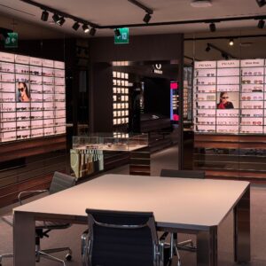 Inside an Oakley store, filming in on location. There are lots of brightly lit glasses boxes where the glasses for each brand sit. There is a table with chairs in the middle of the room.